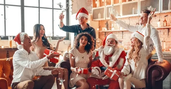 Hosting holiday gatherings in a cozy loft apartment with exposed brick walls, featuring a group of friends celebrating Christmas together on a leather sofa, exchanging wrapped gifts, wearing Santa hats and festive attire, with champagne glasses raised, confetti falling, and a coffee table spread with snacks, drinks, and candles creating a warm intimate party atmosphere.