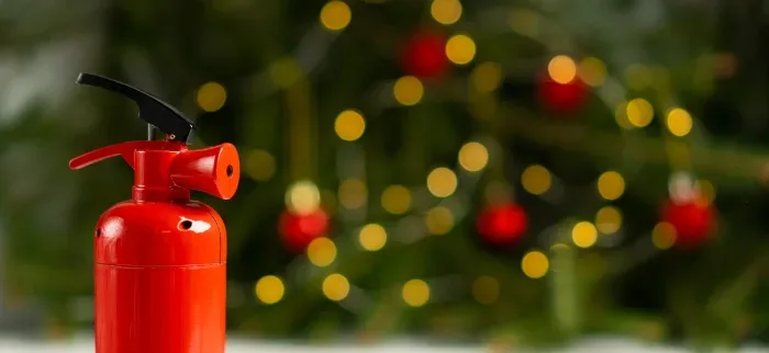 Red fire extinguisher on white surface with decorated Christmas tree, twinkling lights, and red ornaments in soft focus background &ndash; Keep Your Home Safe This Christmas with proper fire safety equipment during the holiday season