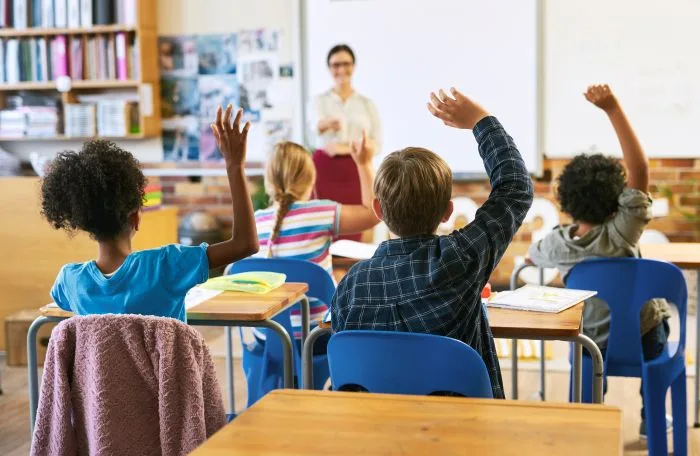Elementary students raise their hands in a sunlit classroom while a smiling teacher stands at the whiteboard, illustrating a guide to the best local schools for home buyers